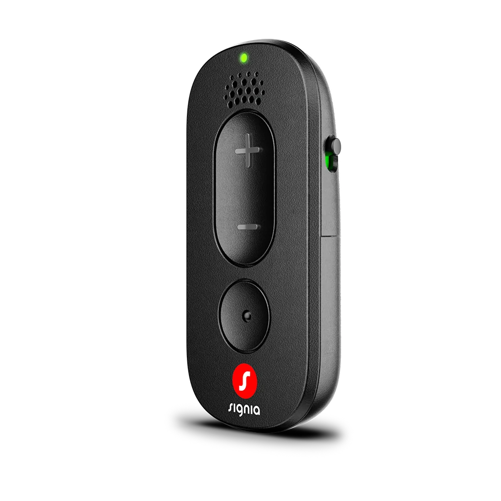 A compact Signia Remote Control in black, equipped with volume buttons, a prominent central button, a speaker grille, and a glowing green indicator on the side, standing upright against a light gray background.